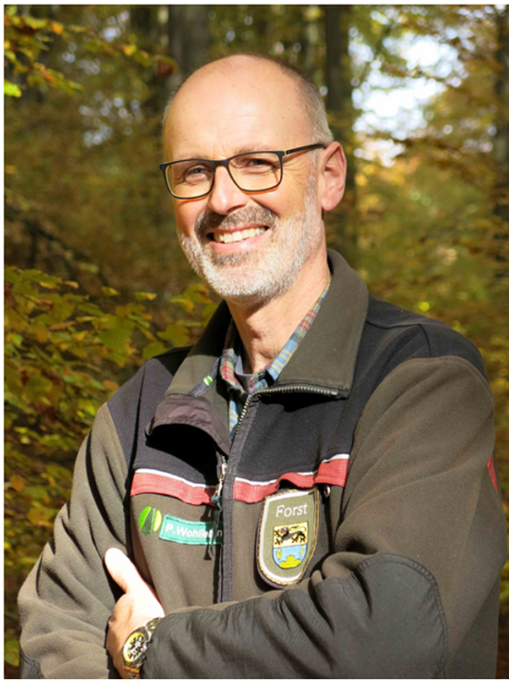 a man stands smiling with arms crossed in a forest